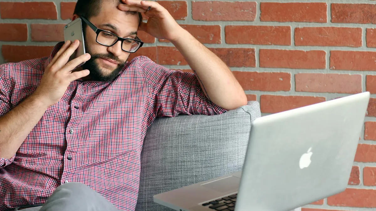 Bearded man looks frustrated while working remotely with laptop and phone.