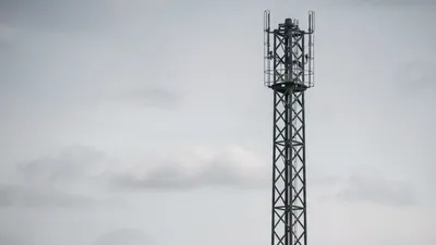 A tall, dark grey mobile phone mast with a lattice structure stands against a cloudy, light grey sky.