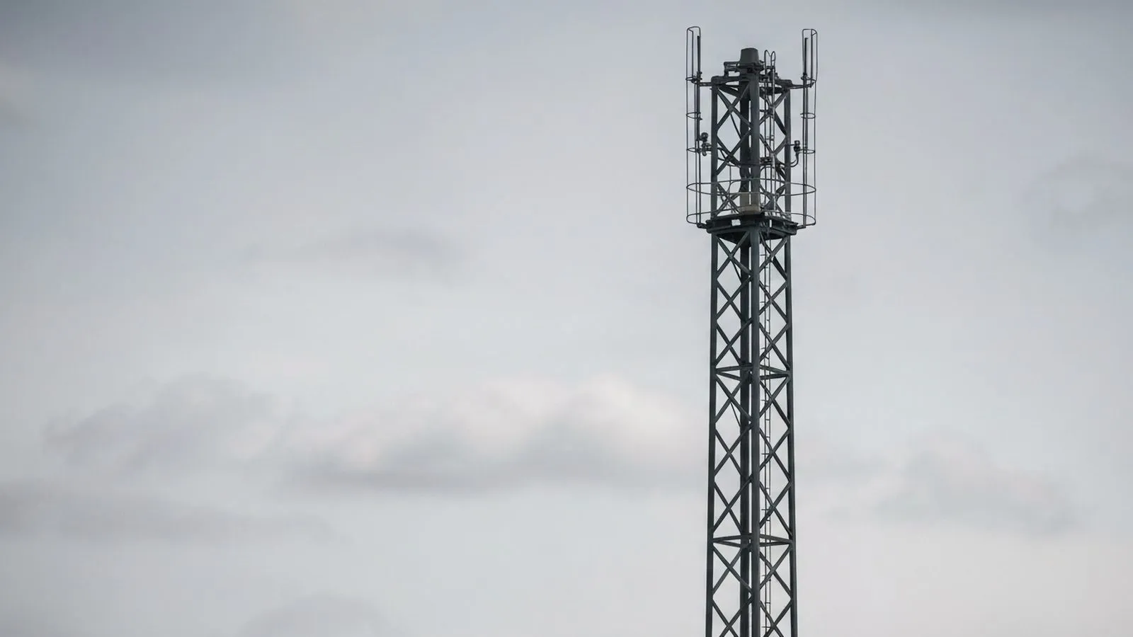 A tall, dark grey mobile phone mast with a lattice structure stands against a cloudy, light grey sky.
