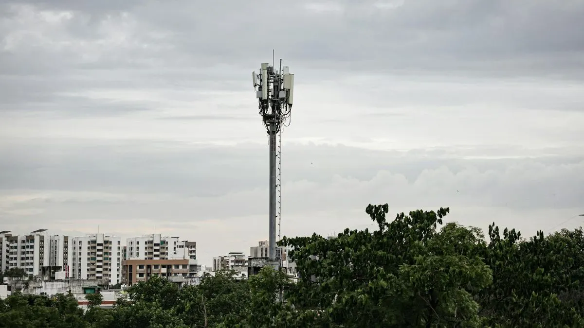 A tall O2 mast stands prominently against a cloudy sky, surrounded by trees and buildings.