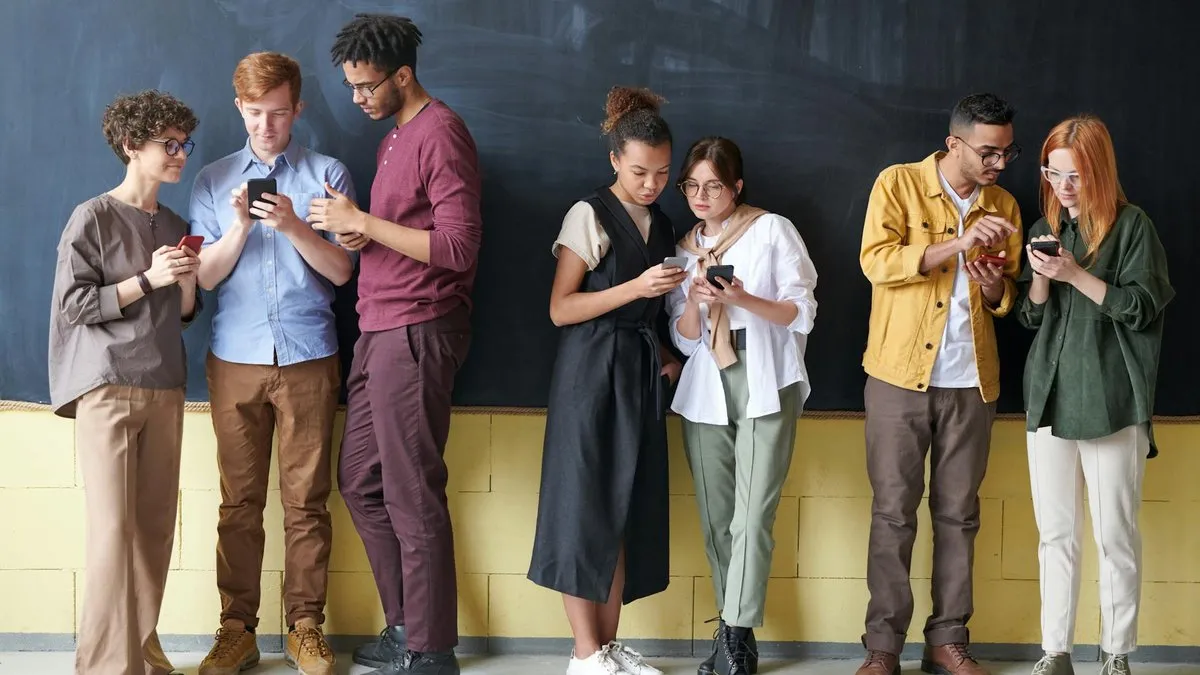 A diverse group of seven young adults standing against a dark wall, all looking at their phones.