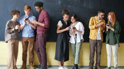 A diverse group of seven young adults standing against a dark wall, all looking at their phones.