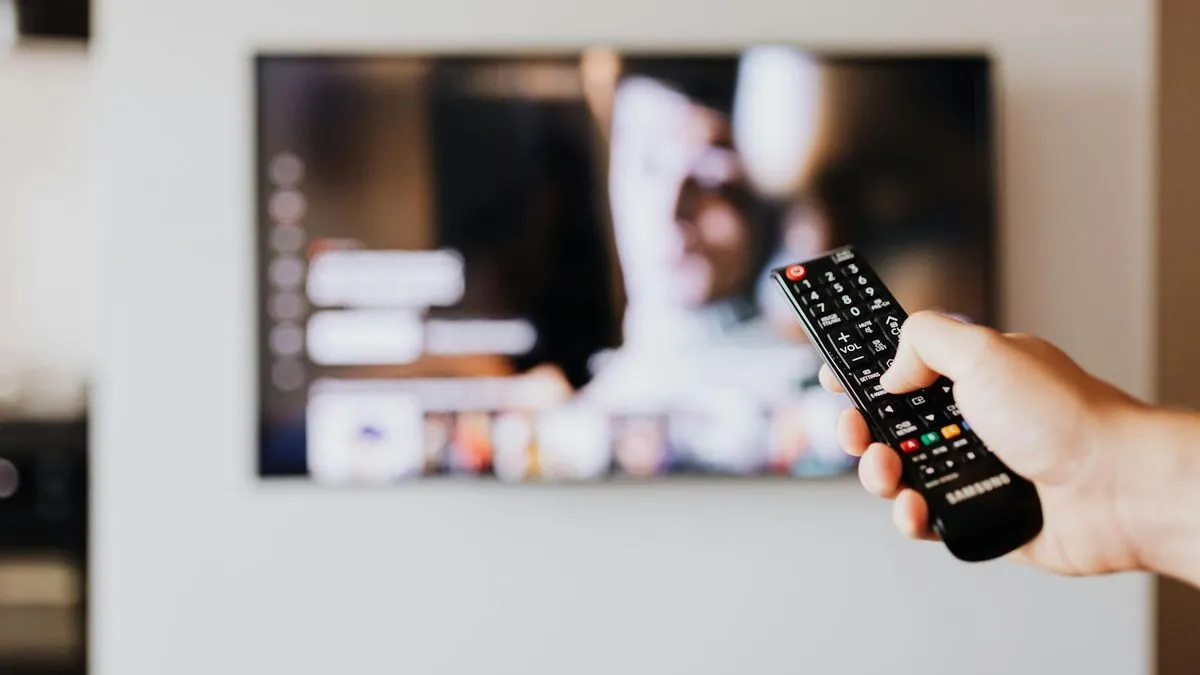 A hand holding a black TV remote control, pointing towards a blurred modern TV screen.