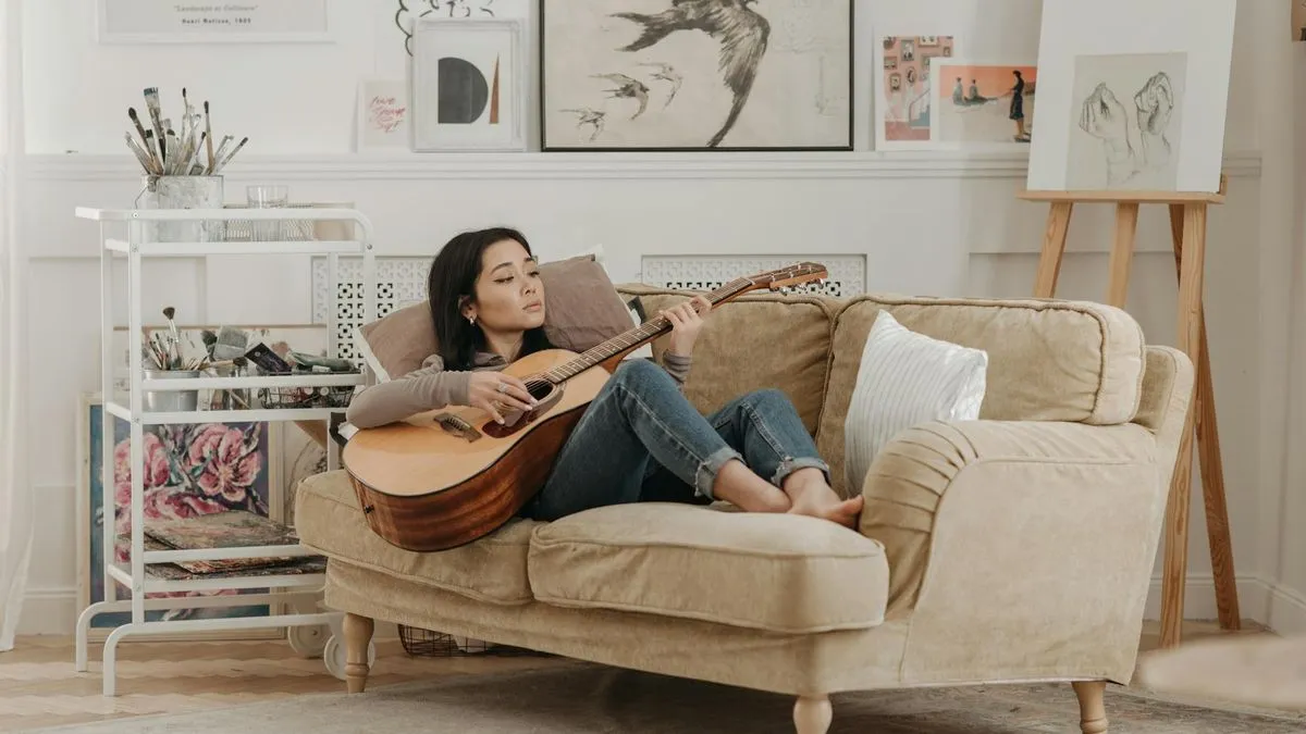 A woman playing an acoustic guitar while relaxing on a beige sofa in a bright living room.