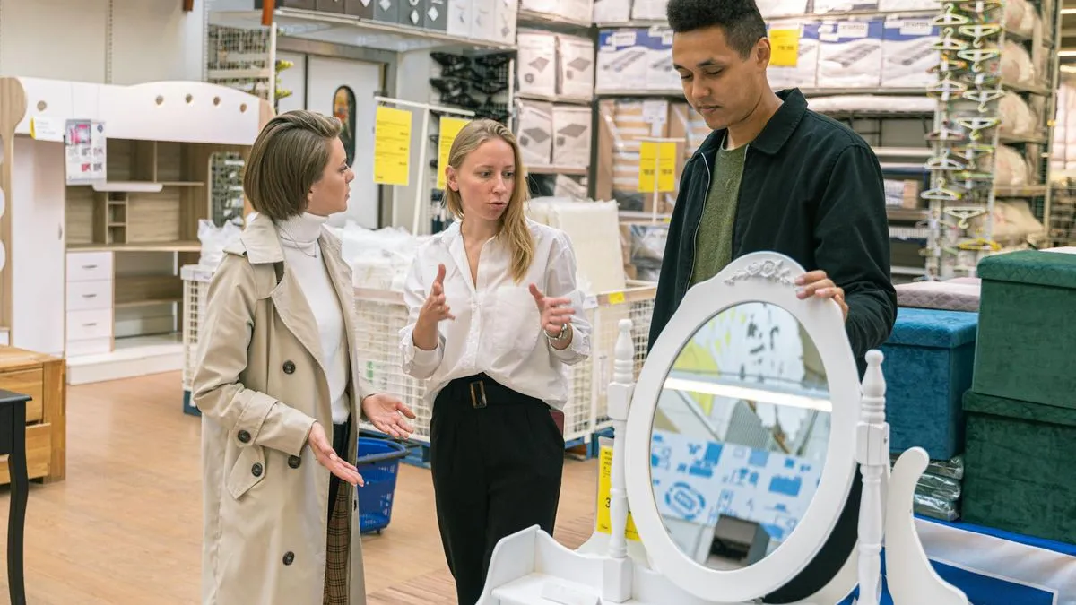 Three people discussing furniture in a store, with a white ornate mirror in the foreground.