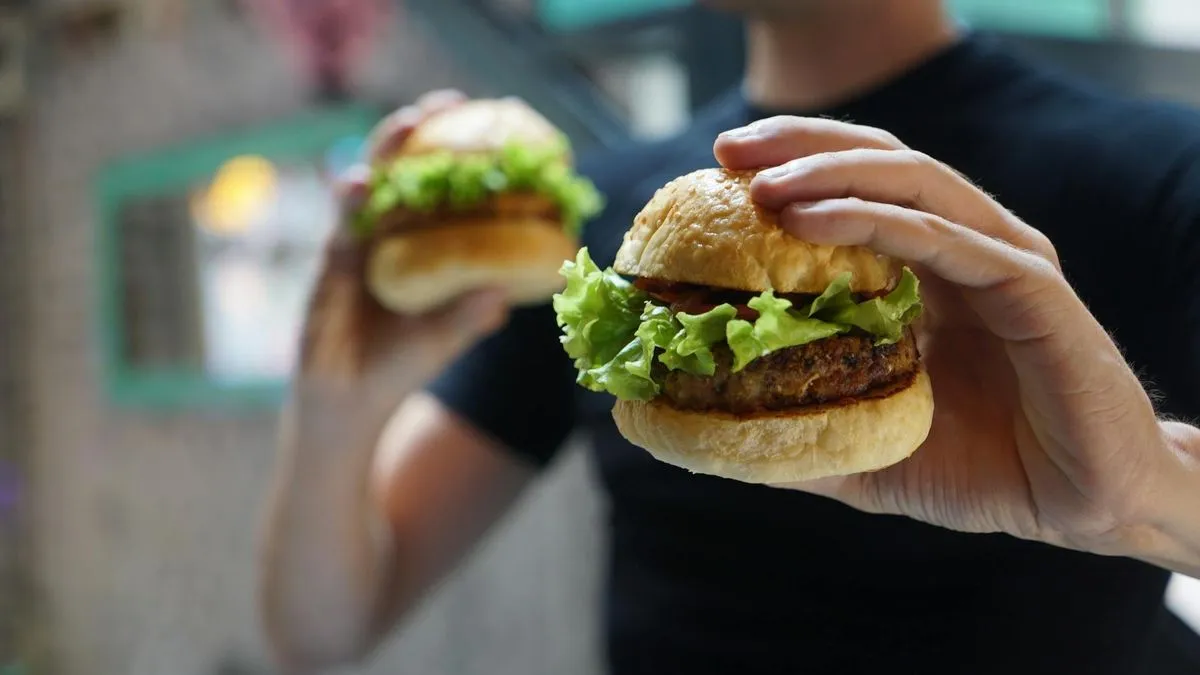 A person holds two fresh burgers with lettuce and meat patties in their hands.