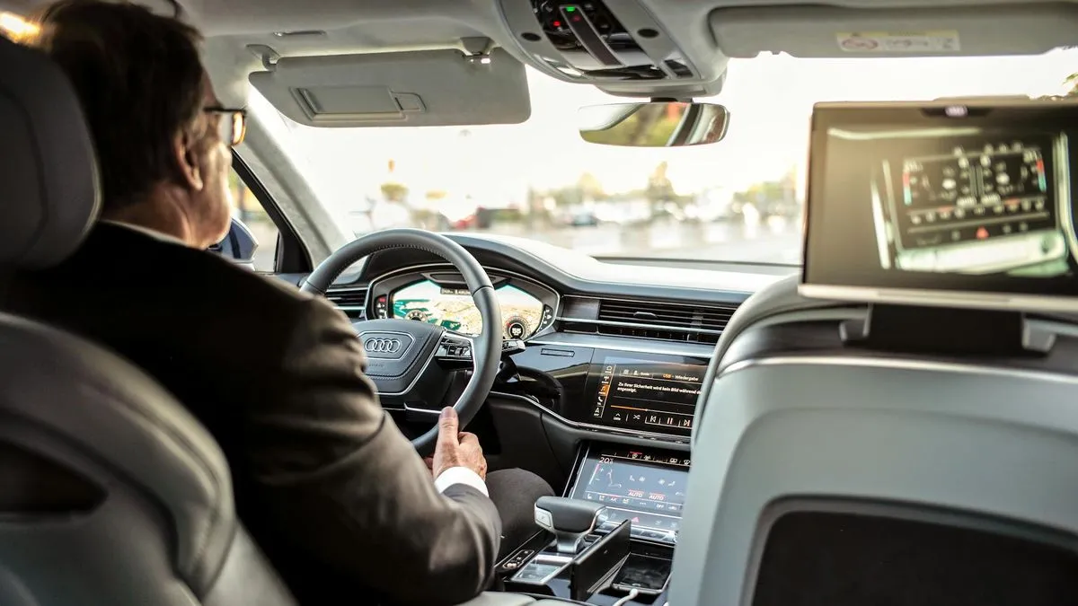 A man in a suit driving an Audi, with a screen displaying car controls in the back.