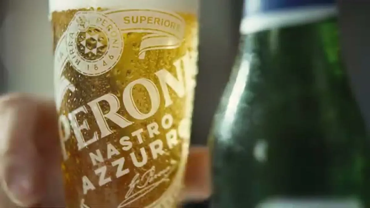 A close-up of a Peroni Nastro Azzurro beer glass, filled with golden beer and foam, next to a green bottle.