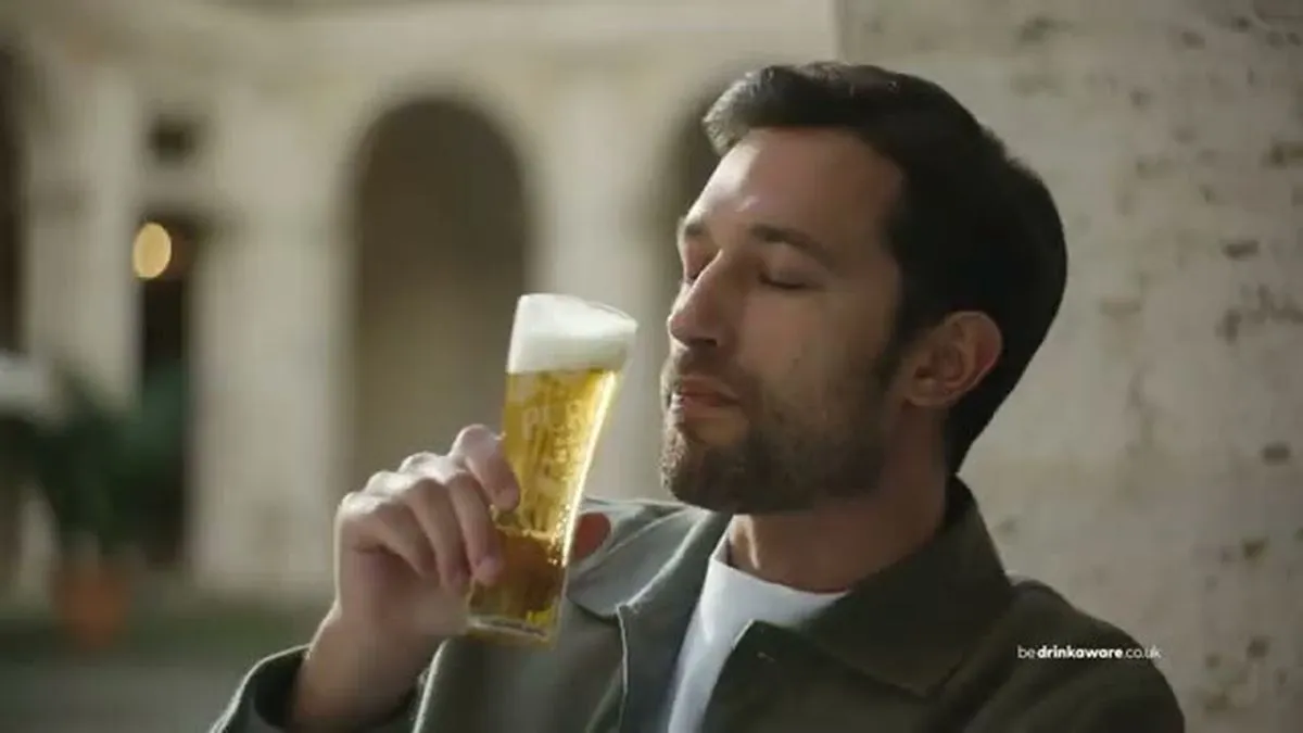 A man with a beard and dark hair, wearing a green jacket, holds a glass of beer, eyes closed, smelling it.