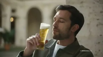 A man with a beard and dark hair, wearing a green jacket, holds a glass of beer, eyes closed, smelling it.