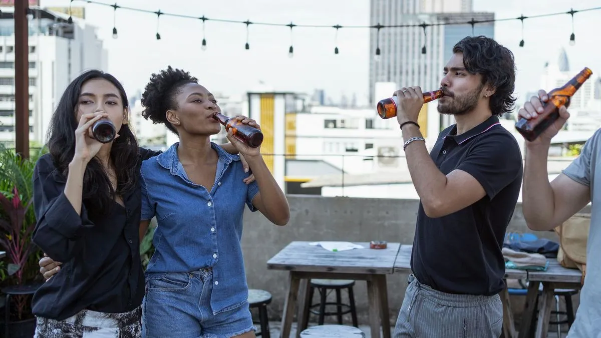 Three friends on a rooftop terrace drinking beer from brown bottles, with city buildings in the background.