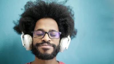 A man with an afro and glasses smiles with his eyes closed, wearing white headphones.