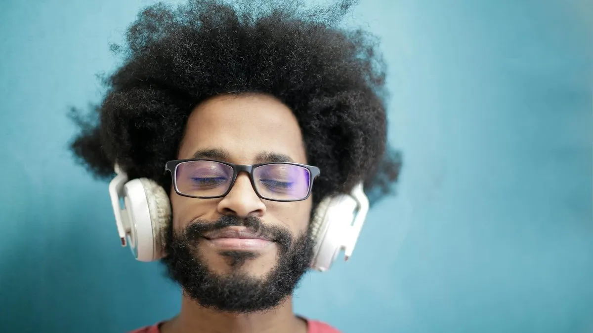 A man with an afro and glasses smiles with his eyes closed, wearing white headphones.