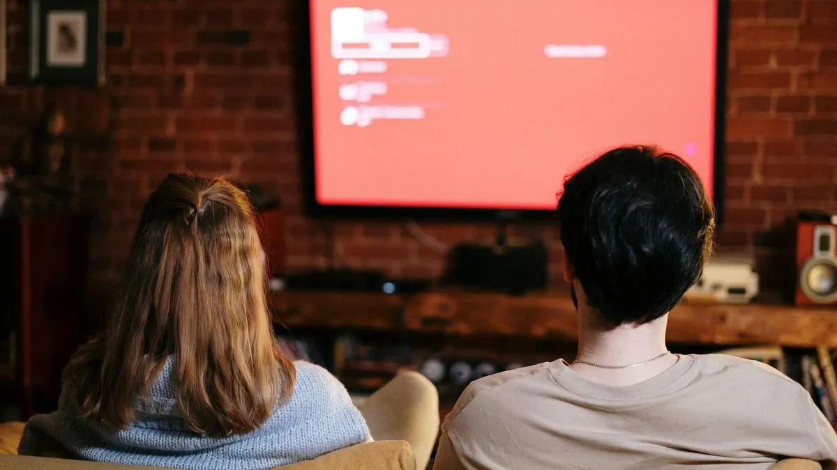 A couple sits on a couch, watching a television screen displaying a red interface.