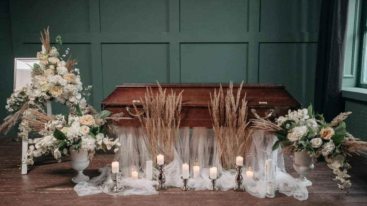 A coffin surrounded by white flowers, candles, and dried grass in a solemn setting.