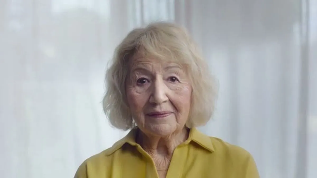 An elderly woman with blonde hair and a yellow shirt looks directly at the camera with a slight smile.