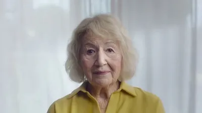 An elderly woman with blonde hair and a yellow shirt looks directly at the camera with a slight smile.