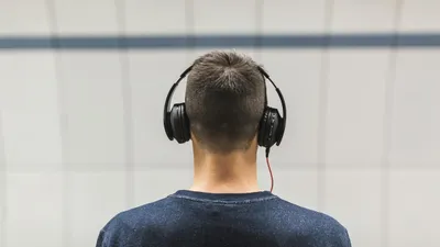 A person from behind wearing black over-ear headphones, listening to music against a plain background.