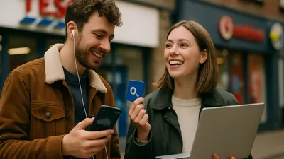 A man with earphones looking at his phone and a woman holding a laptop and a blue card, both smiling.