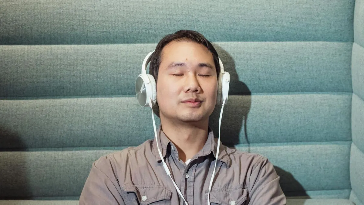A man with closed eyes and white headphones sits against a textured grey wall.
