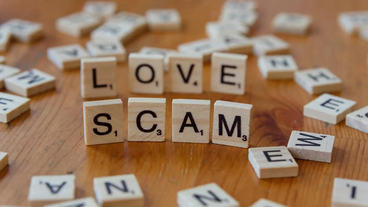 Scrabble tiles form words 'LOVE' and 'SCAM' on a wooden surface, illustrating deceit in relationships.