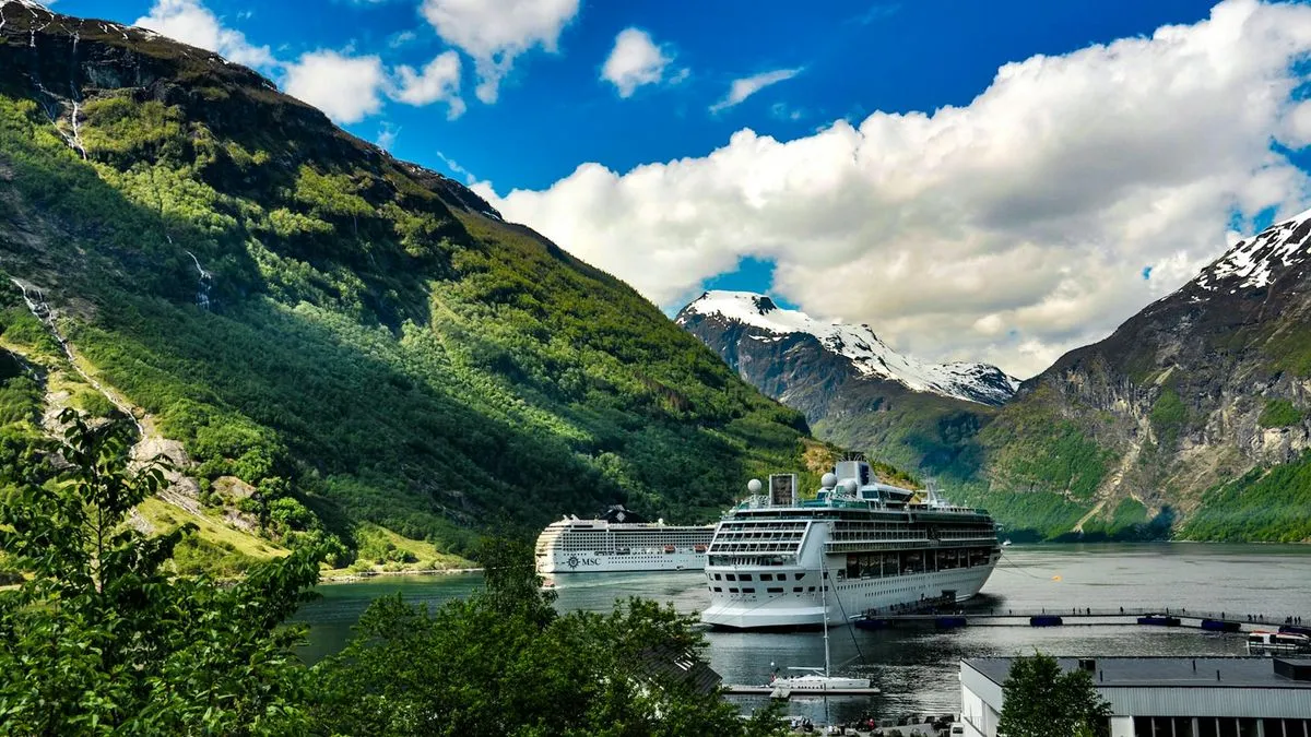 Two large cruise ships docked in a scenic fjord surrounded by lush green and snow-capped mountains.
