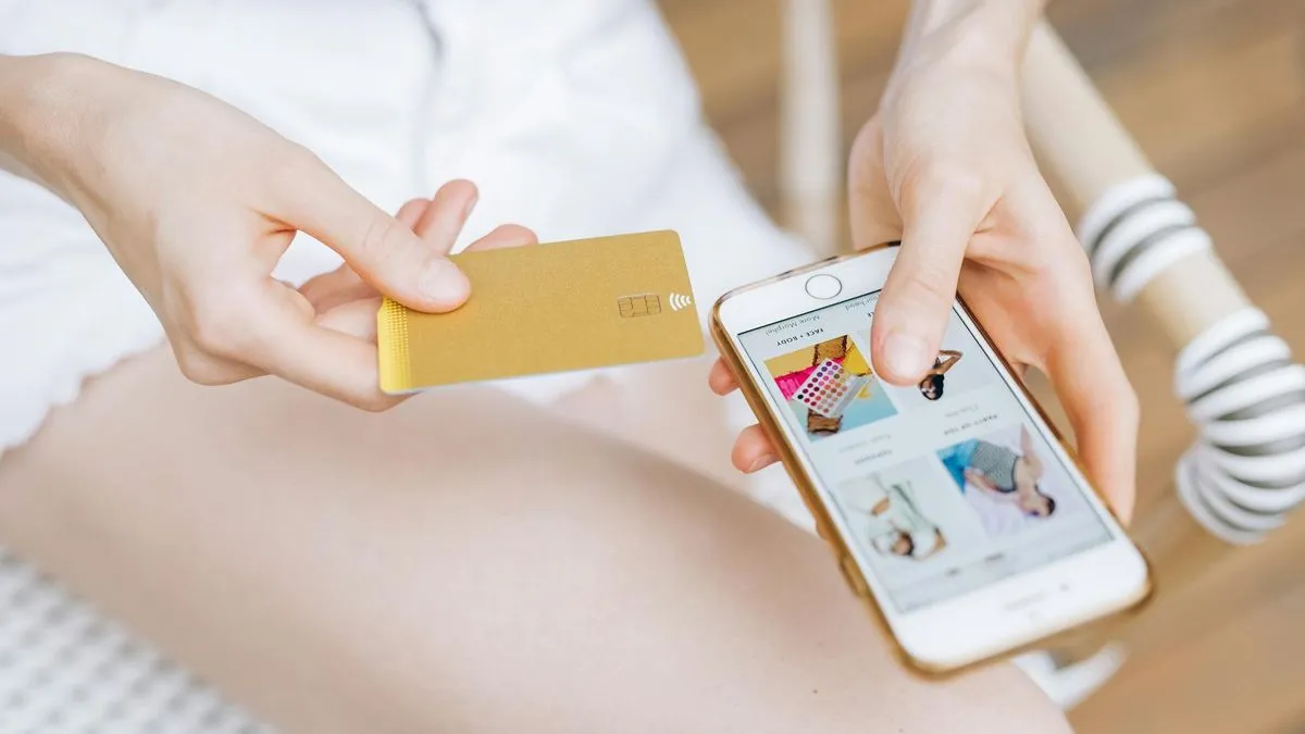 A person holds a gold credit card in one hand and a smartphone displaying an online store in the other.