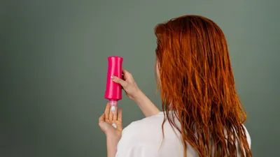 A woman with long red hair squeezes hair product from a pink bottle into her hand.