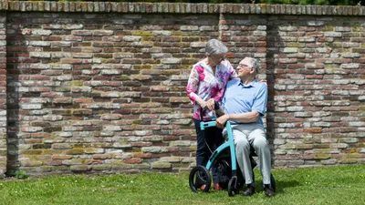 An elderly woman pushing an elderly man in a wheelchair with a brick wall in the background.