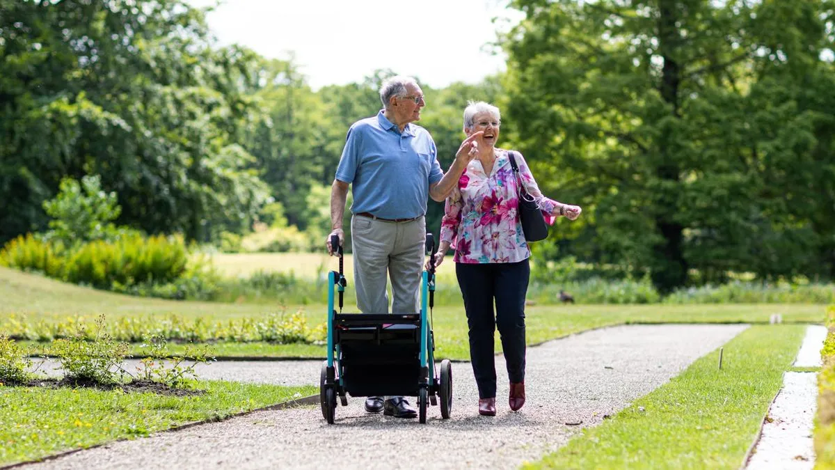 An elderly couple walks on a path, the man using a rollator, the woman smiling.