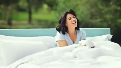 A woman with dark hair smiles, lying in a bed with white sheets outdoors in a green field.