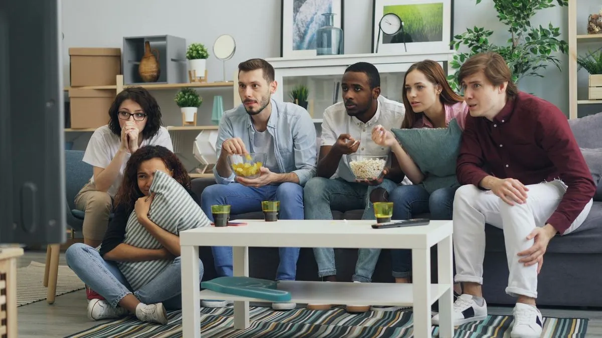 A diverse group of young adults watching TV together in a living room, some eating snacks.