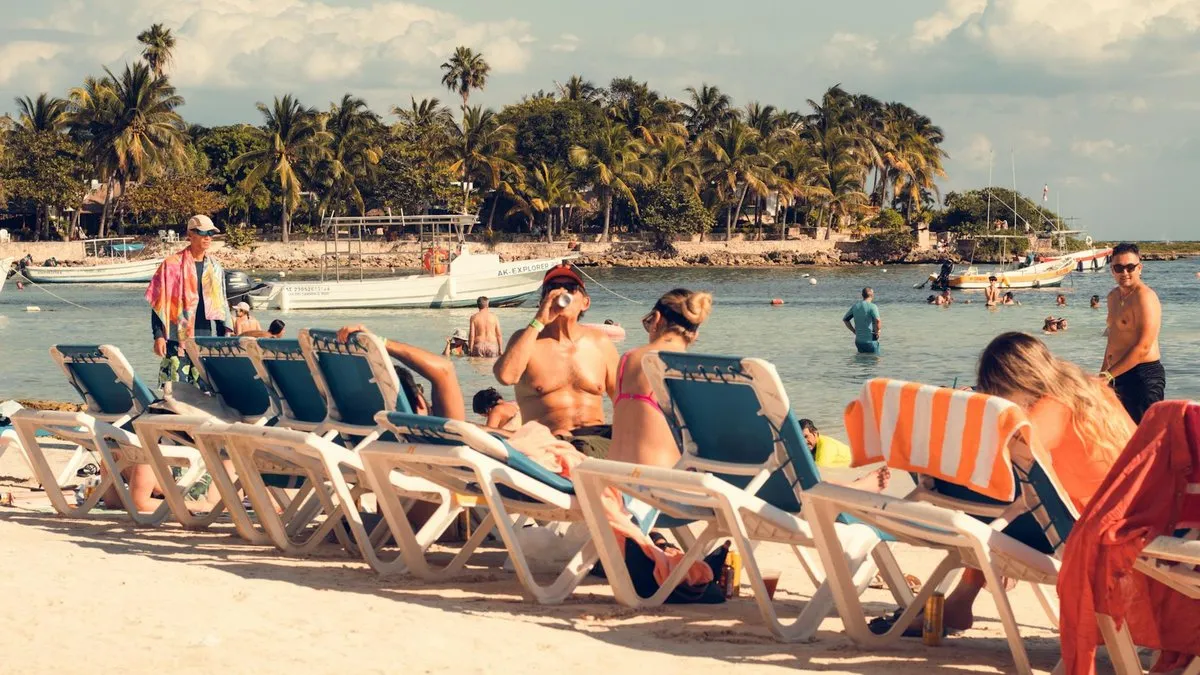 People relaxing on lounge chairs on a sandy beach with palm trees and boats in the background.