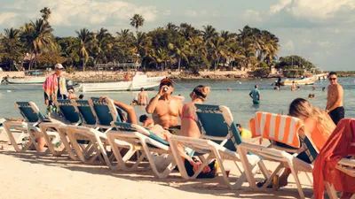 People relaxing on lounge chairs on a sandy beach with palm trees and boats in the background.