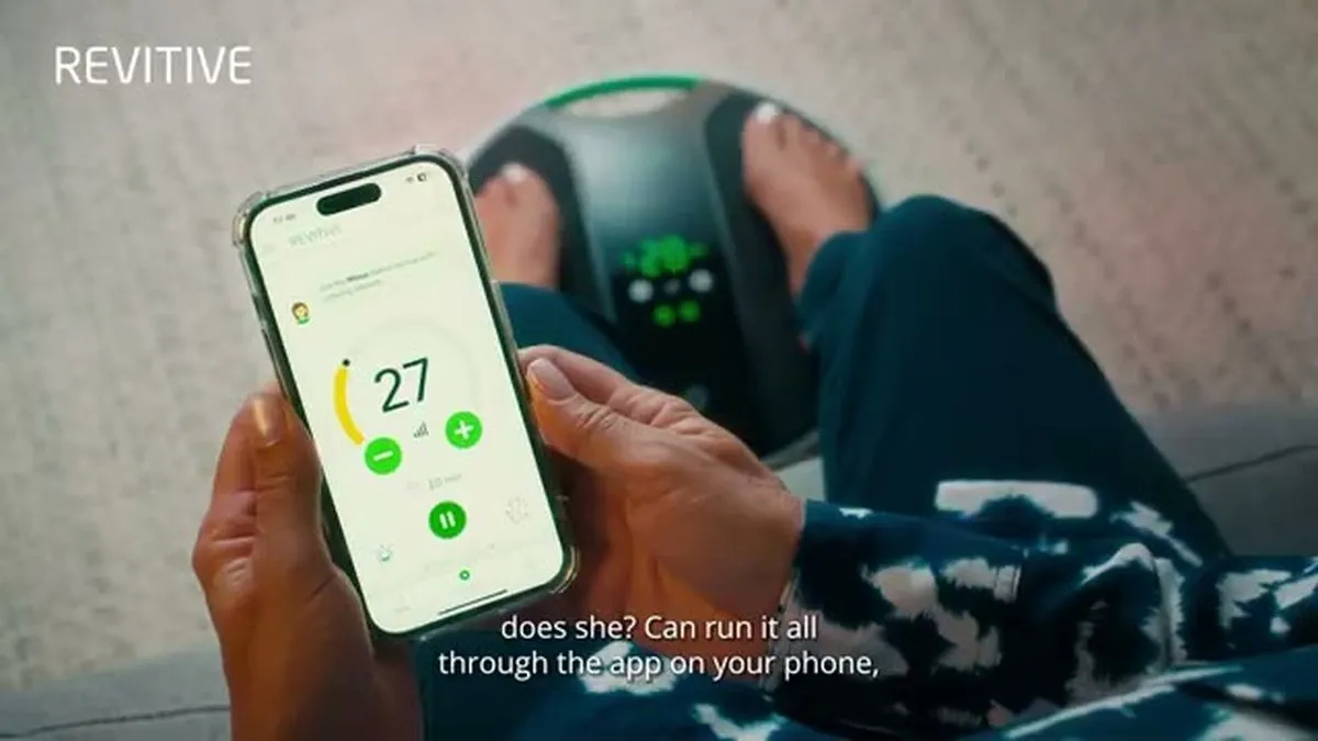 A person's hands hold a smartphone displaying a Revitive app interface, with feet on a Revitive device.
