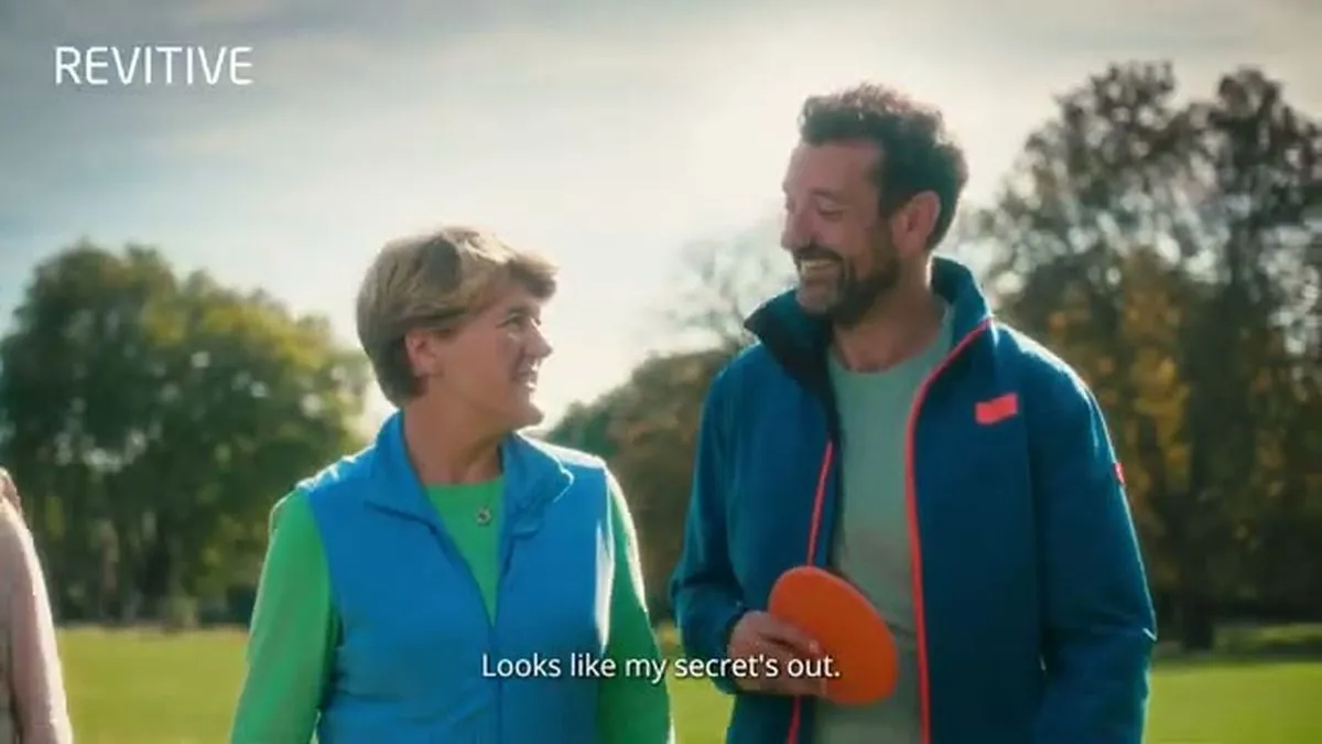 Clare Balding and a man with a beard, both in athletic wear, smile at each other outdoors in a park.