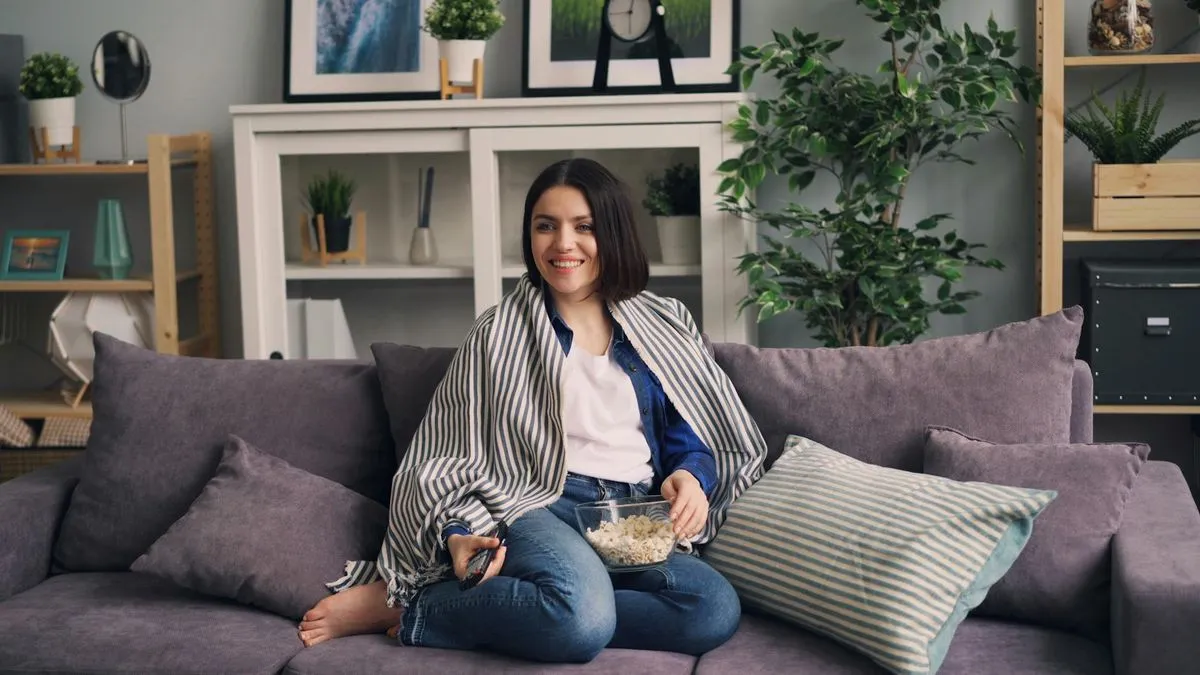 A smiling woman wrapped in a blanket, holding popcorn and a remote, watching TV on a sofa.