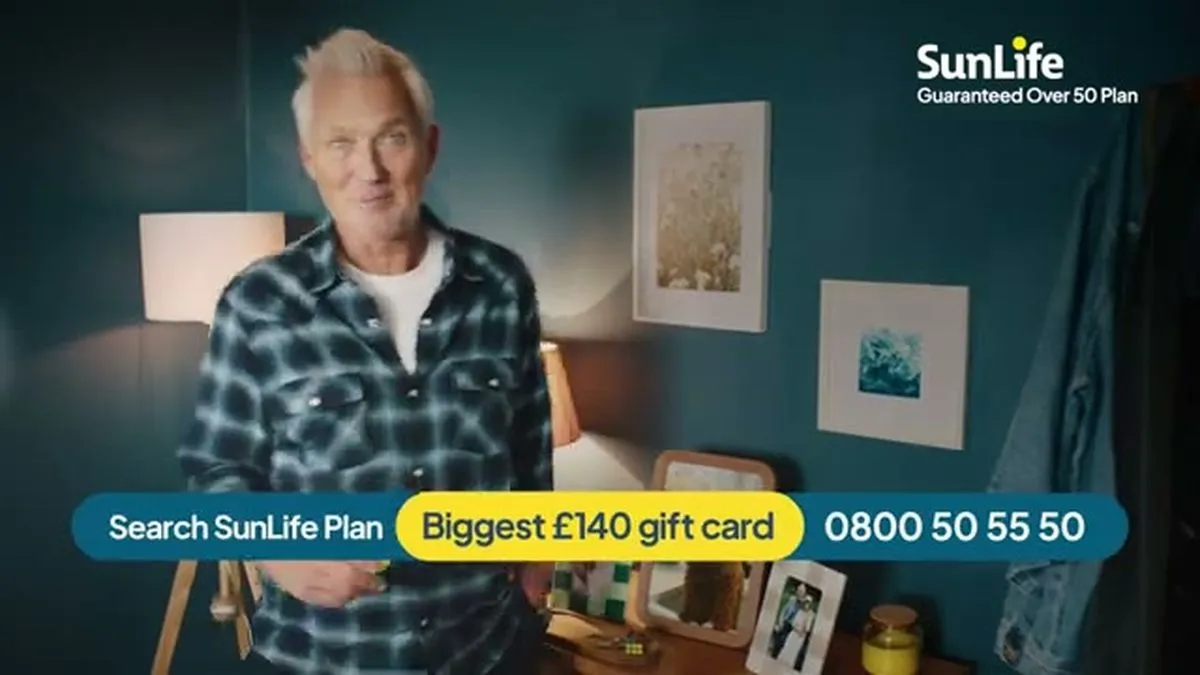 Martin Kemp, a man with white hair, smiles directly at the camera in a medium shot, wearing a blue plaid shirt.