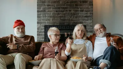 Four older adults sitting on a couch, watching TV and eating snacks.