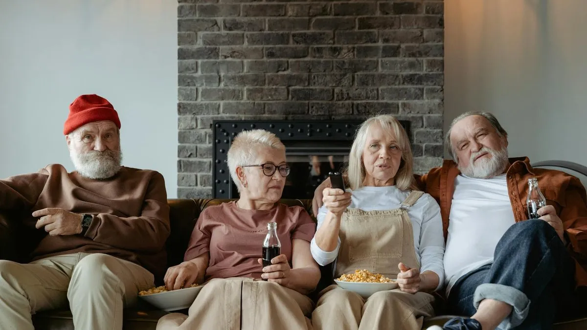 Four older adults sitting on a couch, watching TV and eating snacks.