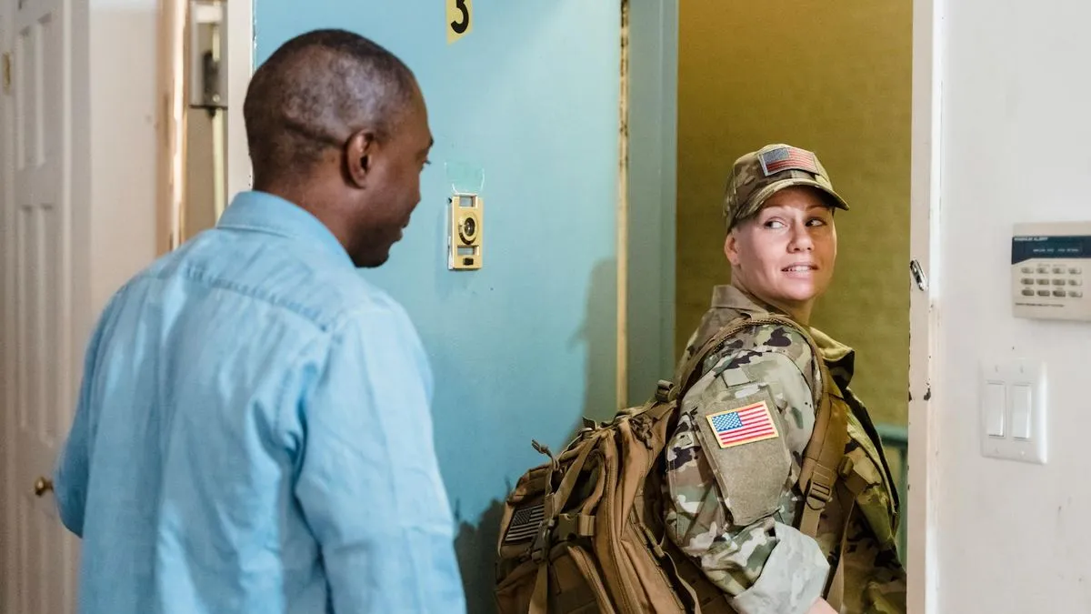 A man watches a woman in military uniform with a backpack enter a doorway.