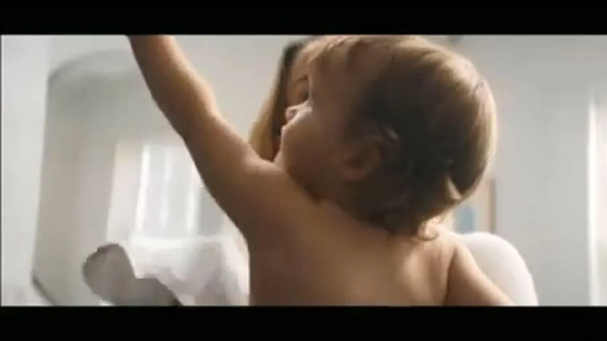 A baby with light brown hair and bare shoulders reaches up, held by an unseen person.