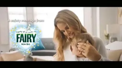 A woman with long blonde hair holds a baby close, both looking down, in a soft-focus living room.