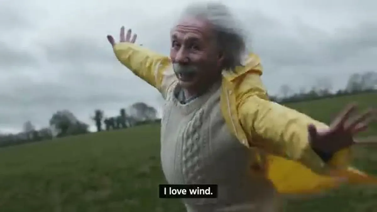An older man with white hair and mustache, wearing a yellow raincoat, smiles with arms outstretched, with 'I love wind' subtitles.