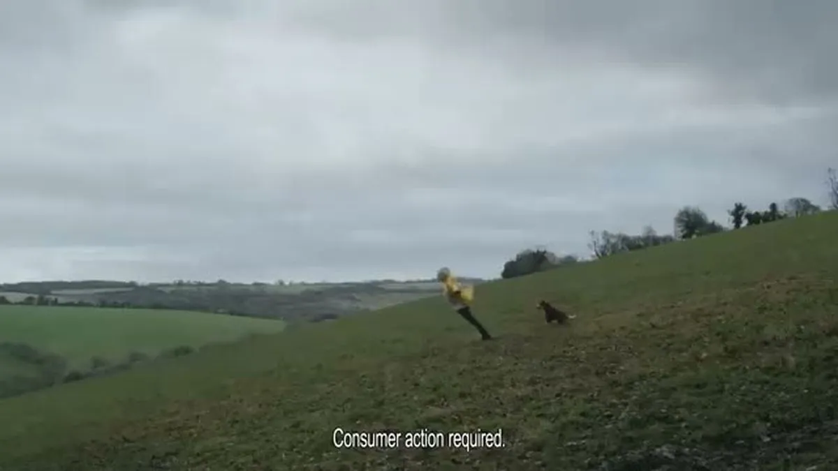 A wide shot of a man in a yellow raincoat and a dog running up a green grassy hill under a cloudy sky.