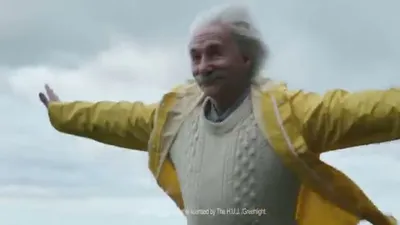 An older man with white hair and mustache, dressed in a yellow raincoat and white sweater, smiles with arms outstretched against a cloudy sky.