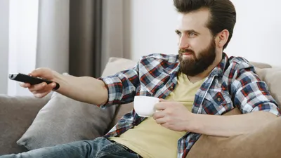 A bearded man in a plaid shirt holding a remote control and a white mug on a sofa.
