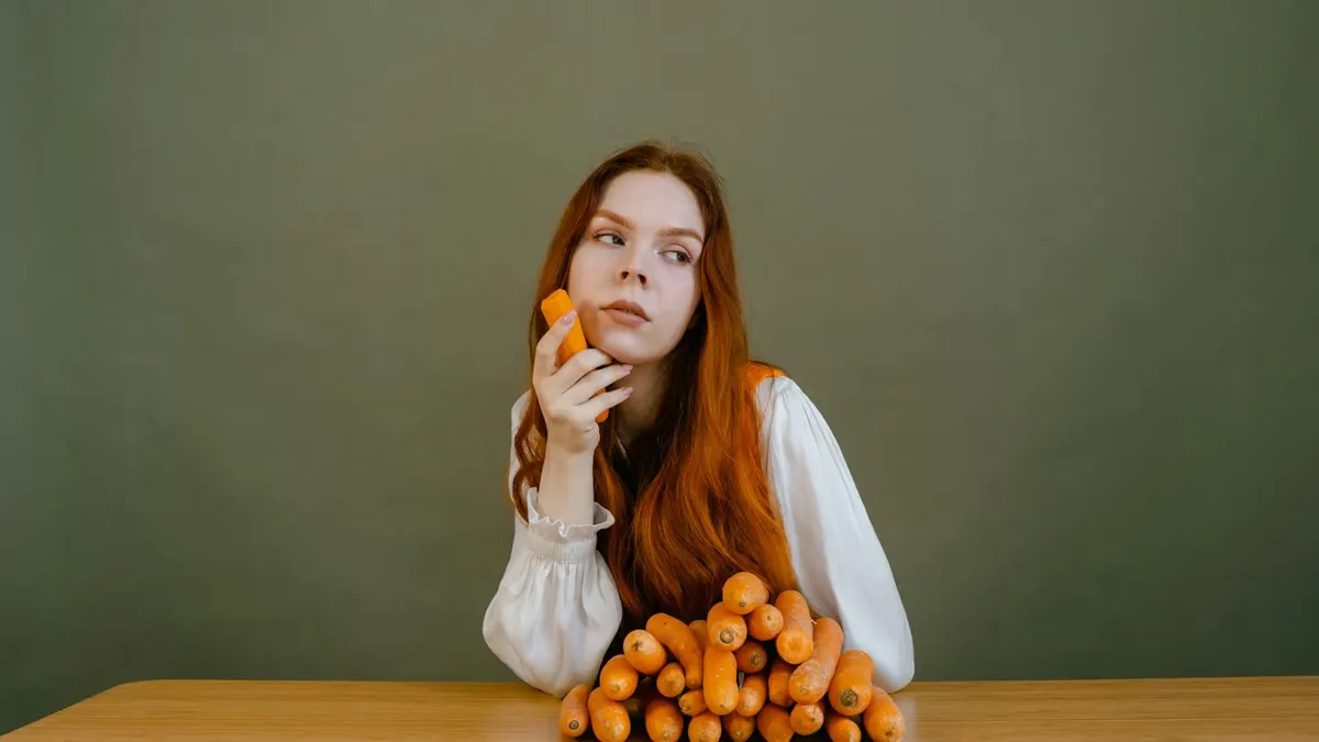 Red-haired woman in white sleeves holding fresh carrots on a wooden table against a green background.