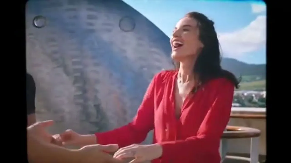 A woman in a red top laughs while talking to someone off-camera on a ship's deck.