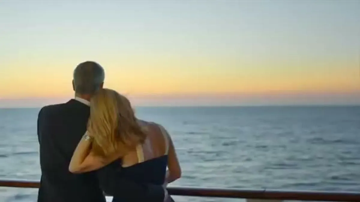 A couple stands with their backs to the camera, looking out at the ocean during sunset from a ship's deck.